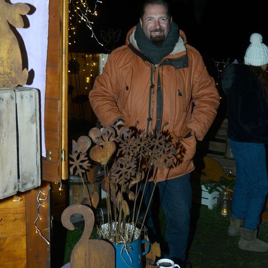 Ein Mann in einer braunen Jacke steht neben einem blauen Eimer mit einem Metallblumenarrangement. Eine Frau in einem weißen Hut und Stiefeln steht im Hintergrund. Die Umgebung ist draußen mit dekorativen Lichtern.