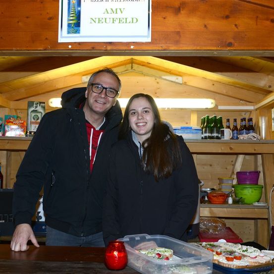 Ein Mann und eine Frau stehen in einem Holzhäuschen, lächeln und posieren für ein Foto. Sie sind von Regalen mit Flaschen und Lebensmitteln umgeben. Das Häuschen hat eine Holzdecke mit einem Schild, das AMV Neufeld steht.