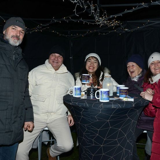 Fünf Personen stehen um einen Tisch mit Tassen, von denen eine eine Tasse hält. Sie lächeln und scheinen eine gesellige Zusammenkunft zu genießen. Der Tisch ist mit einem schwarzen Stoff mit geometrischem Muster bedeckt.