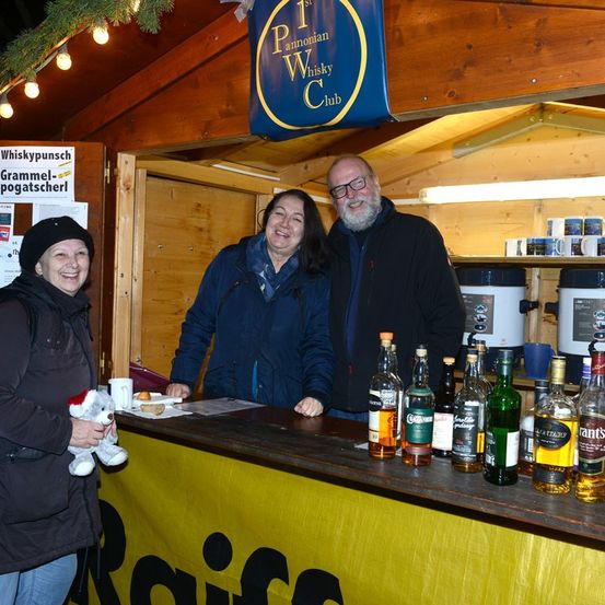 Drei Menschen stehen lächelnd hinter einem Tresen. Der Tresen hat mehrere Flaschen mit Schnaps. Ein Banner über ihnen lautet 1. Pannonischer Whisky Club.