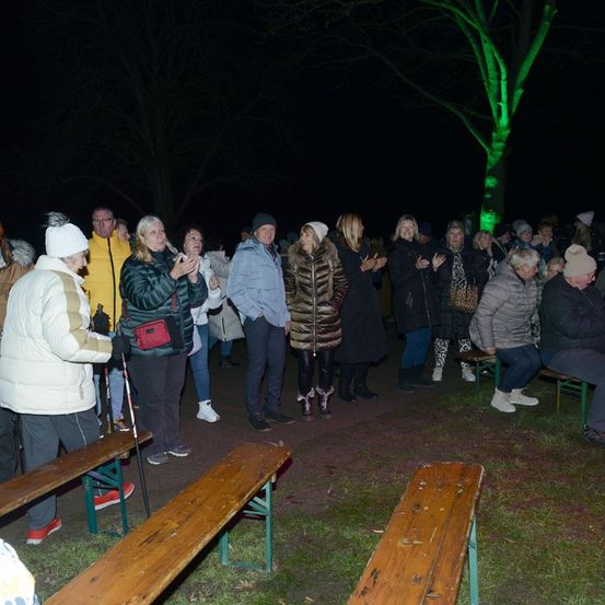Eine Gruppe von Menschen steht im Kreis und klatscht, möglicherweise singen sie. Sie werden von grünen Lichtern eines Baumes in der Mitte beleuchtet. Sie sind warm gekleidet und einige tragen Mützen. Der Ort ist draußen, mit Bänken und Gras.