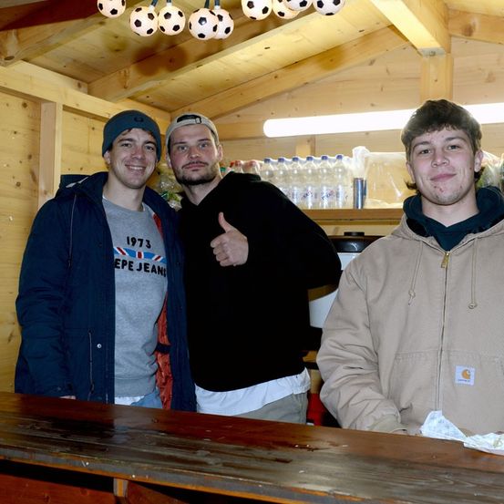Drei junge Männer stehen in einem Holzraum mit einem Holztisch davor. Sie lächeln und scheinen für ein Foto zu posieren. Der Mann in der Mitte gibt einen Daumen hoch. Flaschen und Fußballbälle hängen an der Decke.