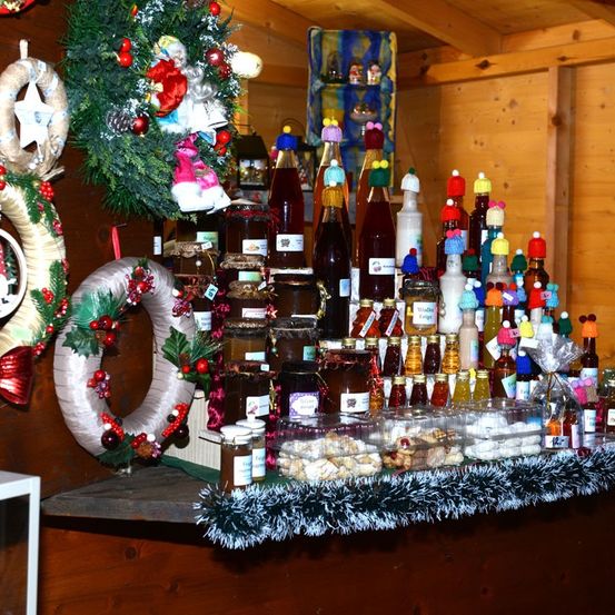 Ein hölzerner Tresen ist mit verschiedenen Weihnachtsartikeln dekoriert, darunter Kränze, Flaschen mit Sirup und Gläser mit Konfitüre. Der Tresen ist mit einer Glitzergirlande geschmückt, und die Regale im Hintergrund sind mit Weihnachtsdekorationen verziert.