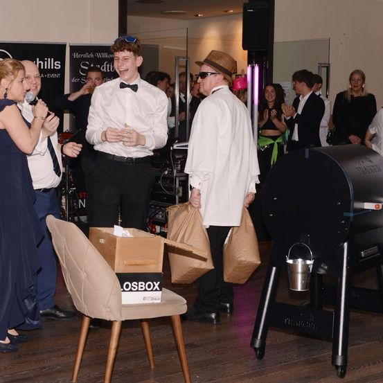 Zwei Männer in formeller Kleidung stehen neben einer Kiste auf einem Stuhl. Sie halten Papiertüten und lächeln eine Frau an, die in ein Mikrofon spricht.