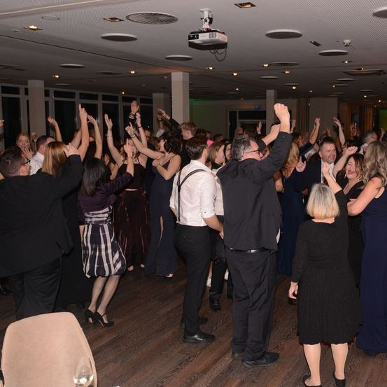 Eine Gruppe von Menschen in formeller Kleidung tanzt in einem Raum mit Holzboden. Einige heben ihre Hände. Ein Beamer hängt von der Decke.
