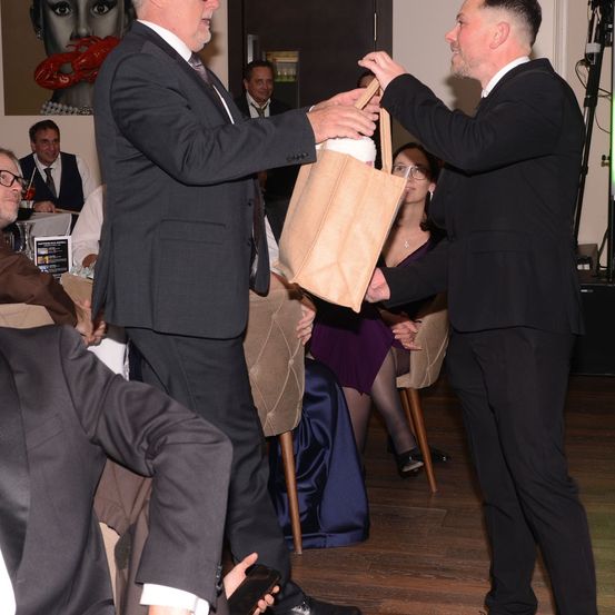 Ein Mann überreicht einem anderen Mann einen braunen Beutel bei einer Veranstaltung. Eine Frau in einem lila Kleid und ein Mann in Anzug und Krawatte sitzen im Hintergrund.