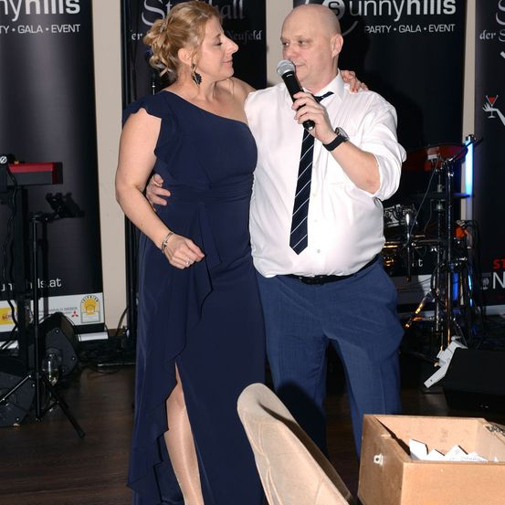 Ein Mann in einem weißen Hemd mit Krawatte hält ein Mikrofon und steht neben einer Frau in einem blauen Kleid. Sie befinden sich in der Nähe einer Bühne mit Musikinstrumenten und einem Hintergrund.