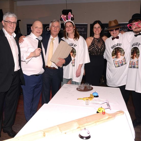 Eine Gruppe von Menschen in Kostümen steht um einen Tisch mit einem Holzstab und einer Glocke darauf. Eine Person hält ein Mikrofon und ein Stück Papier, während eine andere ein Buch in der Hand hat.