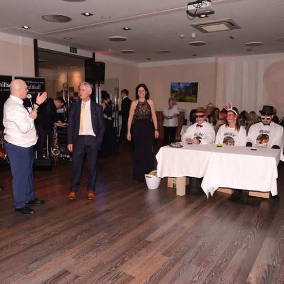 Ein Mann spricht in ein Mikrofon, während ein Mann in Anzug in der Nähe steht. Eine Frau in einem schwarzen Kleid steht hinter einem Tisch mit anderen in Kostümen. Ein Saxophon und ein Lautsprecher sind im Hintergrund.