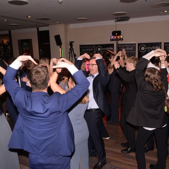 Mehrere Menschen tanzen in einem Raum mit Holzboden. Der Mann in der Mitte trägt eine Fliege und eine Brille. Hinter ihm steht ein Stativ mit einer Kamera. Der Raum ist mit Bannern dekoriert.