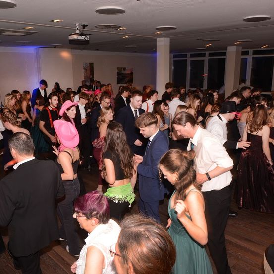 Eine große Menschenmenge in einem Raum, einige tanzen, andere stehen und reden. Einige tragen Hüte und Brillen. Der Raum ist schwach beleuchtet mit blauen Lichtern.