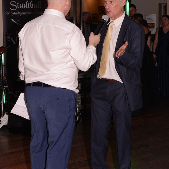 Ein Mann in formeller Kleidung wird von einem anderen Mann in einem formellen Anzug interviewt, beide stehen auf einem Holzboden. Der Interviewer hält ein Mikrofon und gestikuliert während des Sprechens. Im Hintergrund sind mehrere Personen anwesend.