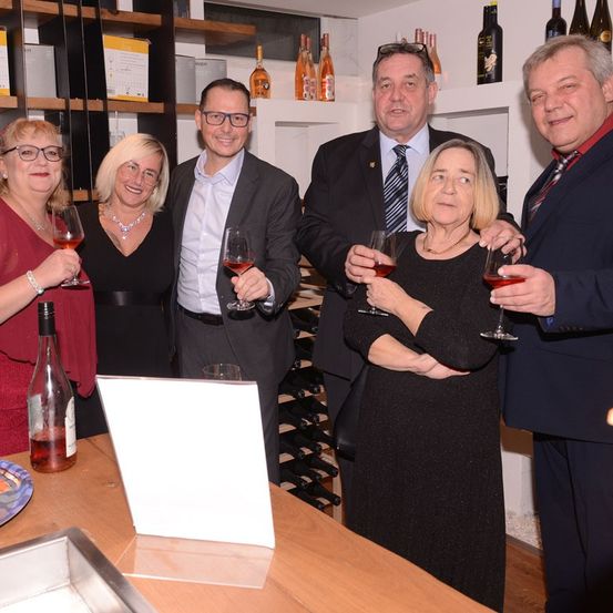 Fünf Erwachsene in formeller Kleidung stehen zusammen in einem Raum, jeder hält ein Weinglas. Sie scheinen ein Ereignis zu feiern. Dahinter sind Regale mit Weinflaschen gefüllt.