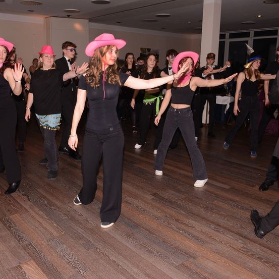 Eine Gruppe von Menschen in schwarzen Kleidern und rosa Hüten tanzt auf einem Holzboden.
