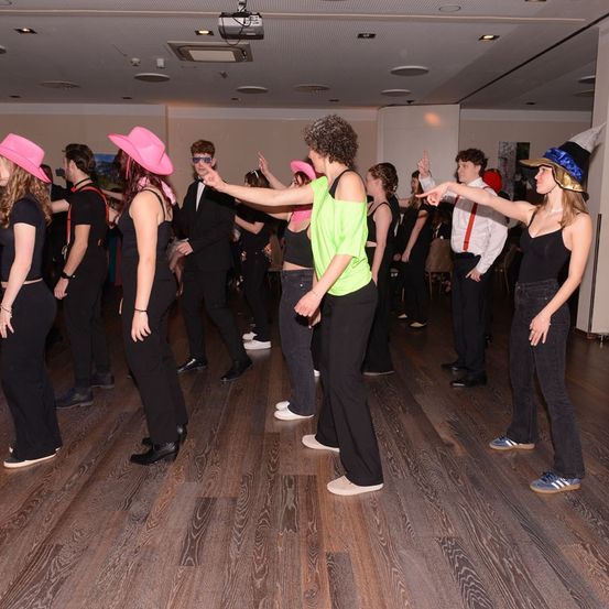 Eine Gruppe von Menschen in einem Raum, die Hüte tragen und tanzen. Einige tragen Schwarz, andere tragen Grün. Der Raum hat einen Holzboden und weiße Wände.