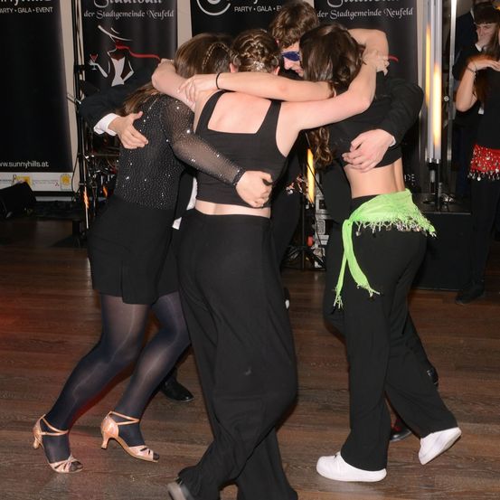 Vier Personen tanzen eng auf einem Holzboden, zwei Paare. Die Frauen tragen schwarze Kleider und hohe Schuhe, während die Männer schwarze Anzüge tragen. Im Hintergrund befinden sich Poster und ein Lautsprecher.