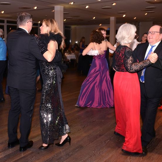 Mehrere Menschen tanzen bei einer formellen Veranstaltung, darunter ein Paar in Schwarz und ein Paar in Rot und Lila. Der Boden ist aus Holz und der Raum ist schwach beleuchtet.