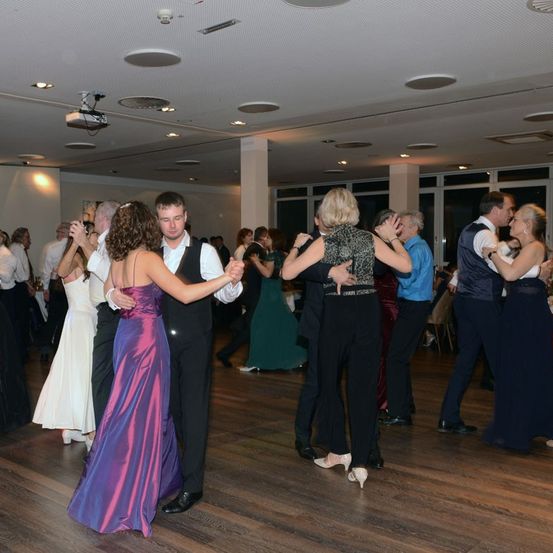 Eine Gruppe von Menschen in formeller Kleidung tanzt auf einer hölzernen Tanzfläche. Sie sind in verschiedenen Posen und einige halten sich an den Händen. Eine Projektor ist an der Decke montiert.