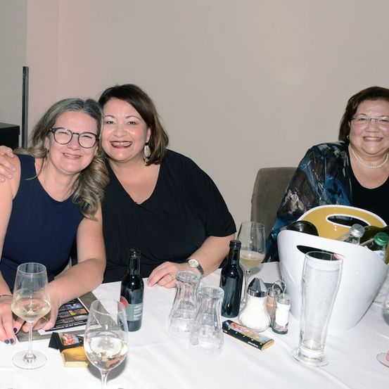 Drei Frauen sitzen am Tisch und lächeln. Eine hat ihren Arm um die andere gelegt, vor ihnen stehen Weinflaschen und Gläser. Ein Eimer mit Flaschen steht ebenfalls auf dem Tisch.