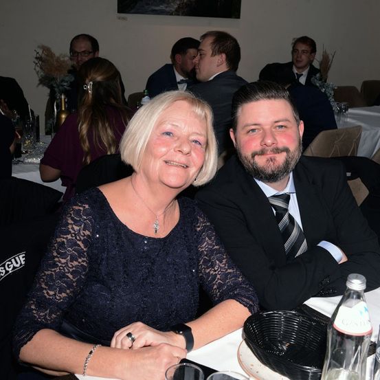 Eine Frau mit blonden Haaren sitzt neben einem Mann in Anzug bei einer formellen Veranstaltung. Sie lächeln beide. Es sitzen noch andere Leute am Tisch. Flaschen, Gläser und ein Korb stehen auf dem Tisch.