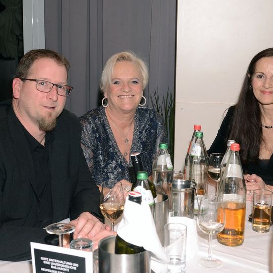 Drei Personen sitzen an einem Tisch, lächeln und posieren für ein Foto. Eine Tischdekoration umfasst Weingläser, Flaschen und eine schwarze Karte mit Text.