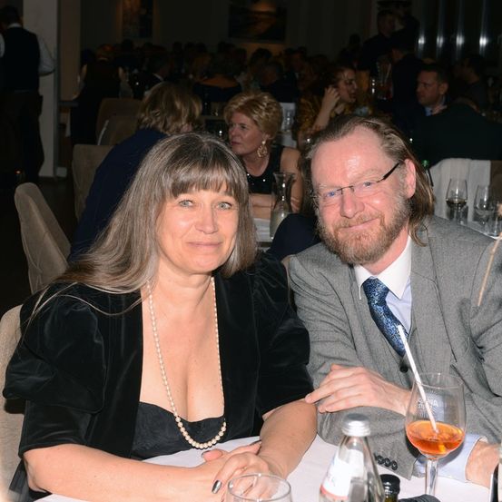 Ein Mann und eine Frau sitzen bei einer formellen Veranstaltung zusammen. Die Frau trägt ein schwarzes Kleid und eine Perlenkette, während der Mann einen Anzug und eine Brille trägt. Hinter ihnen sitzen andere Gäste an Tischen und unterhalten sich.