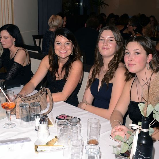 Drei Frauen sitzen an einem Tisch und lächeln für ein Foto. Die Frau links hat ein Getränk und eine Handtasche. Vor ihnen stehen Gläser, eine Flasche und eine Speisekarte. Im Hintergrund sind andere Menschen zu sehen.