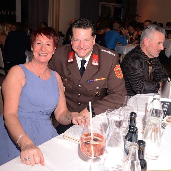 Eine Frau in einem blauen Kleid und ein Mann in Uniform sitzen an einem Tisch und lächeln. Vor ihnen stehen ein Glas Wein, eine Flasche und ein Buch.