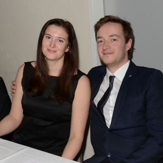 Ein Mann und eine Frau sitzen nebeneinander an einem Tisch, lächeln und posieren für ein Foto. Die Frau trägt ein schwarzes Kleid und der Mann ist in Anzug und Krawatte.