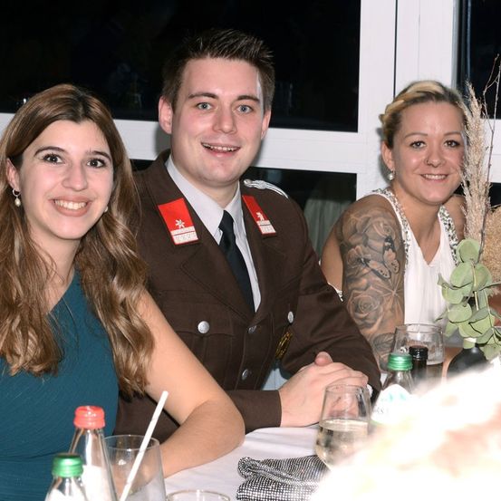 Drei Personen sitzen an einem Tisch und lächeln für ein Foto. Ein Mann in Uniform mit einem roten Abzeichen sitzt zwischen zwei Frauen. Sie haben Gläser und eine Pflanze auf dem Tisch.