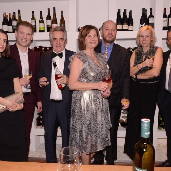 Eine Gruppe von Menschen in formeller Kleidung steht und posiert für ein Foto in einem Weinkeller mit Weinflaschen an den Regalen.
