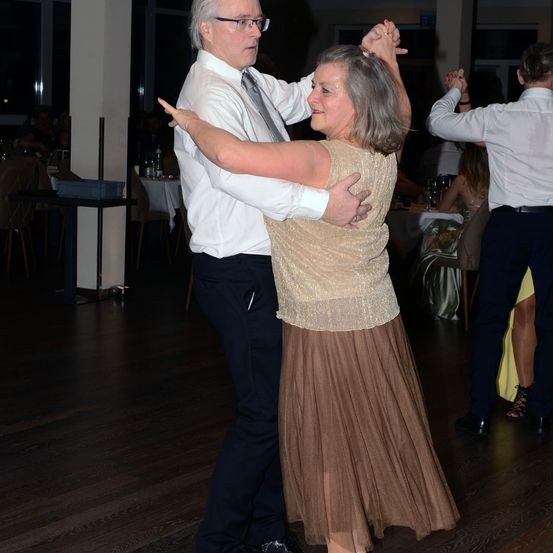 Ein älterer Mann und eine ältere Frau tanzen auf einem Holzboden, lächelnd und Händchen haltend. Ein weiteres Paar tanzt hinter ihnen. Tische mit Flaschen und einer Stange sind im Hintergrund.