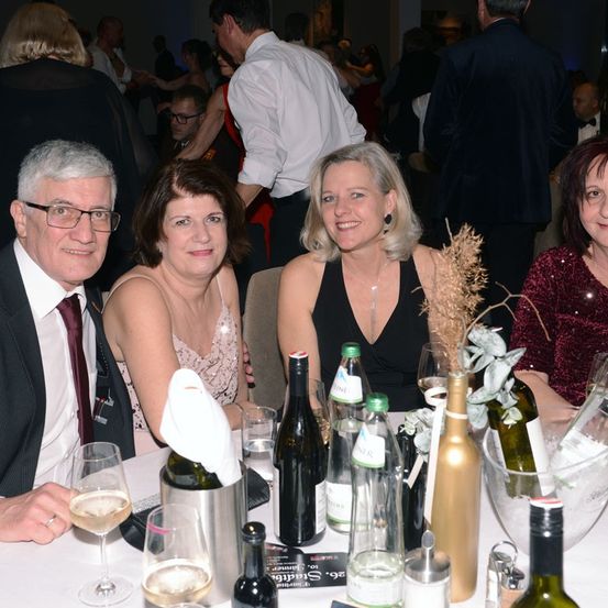 Vier Erwachsene sitzen an einem Tisch mit Weinflaschen und Gläsern in einer gesellschaftlichen Umgebung, möglicherweise einer Feier. Die Frau in der Mitte trägt eine Halskette und lächelt. Der Mann links trägt eine Brille.