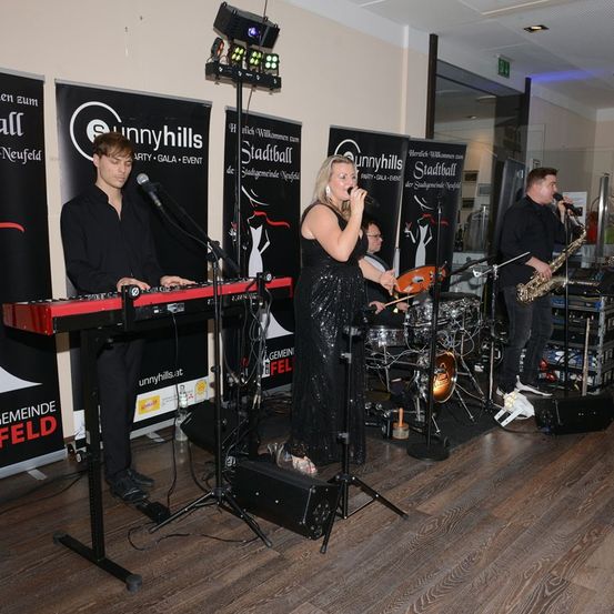 Eine Band tritt auf einer Bühne auf, eine Frau singt und ein Mann spielt Saxophon. Hinter ihnen spielt ein Mann Keyboard. Die Bühne ist von Werbebannern umgeben.