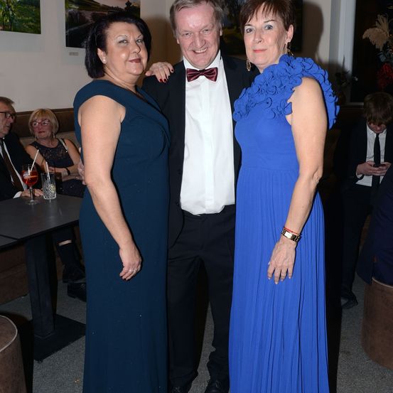 Drei Menschen stehen bei einer Veranstaltung zusammen. Zwei Frauen und ein Mann, wobei der Mann einen schwarzen Anzug und eine Fliege trägt. Die Frauen tragen blaue Kleider, eines ärmellos und das andere mit einem rüschenbesetzten Ausschnitt. Sie befinden sich in einem Raum mit einem Tisch und Stühlen.