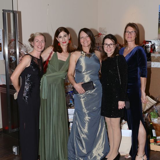 Fünf Frauen in Abendkleidern stehen in einem Raum zusammen. Sie lächeln und scheinen für ein Foto zu posieren. Die Frauen tragen Kleider in verschiedenen Farben. Hinter ihnen befinden sich Gegenstände, die wie Geschenke oder Dekorationen aussehen.