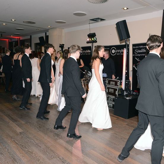 Eine Gruppe formell gekleideter Personen tanzt auf einem Holzboden. Die Braut im weißen Kleid ist von Gästen umgeben. In der Nähe sind Geräte wie Lautsprecher und ein Schlagzeug aufgebaut.