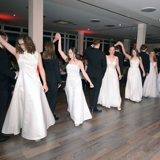 Eine Gruppe von Menschen in formeller Kleidung tanzt in einem Raum, zwei Paare halten Händchen, Frauen in weißen Kleidern.