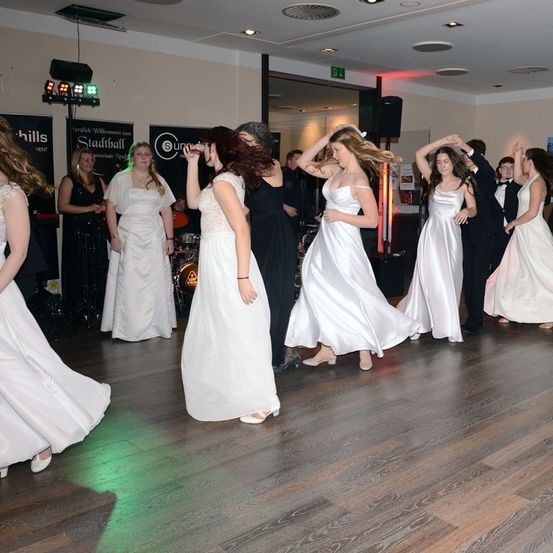 Eine Gruppe von Frauen in weißen Kleidern tanzt auf einem Holzboden, beleuchtet von bunten Lichtern, während andere stehen, möglicherweise bei einer Hochzeitsfeier.