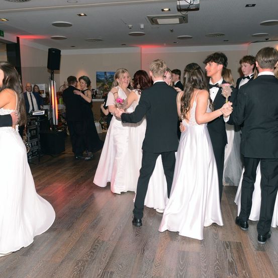 Eine Gruppe von Menschen, wahrscheinlich auf einer Hochzeit, tanzt auf einem Holzboden. Einige halten Blumen, und der Raum hat eine Disco-Beleuchtung.