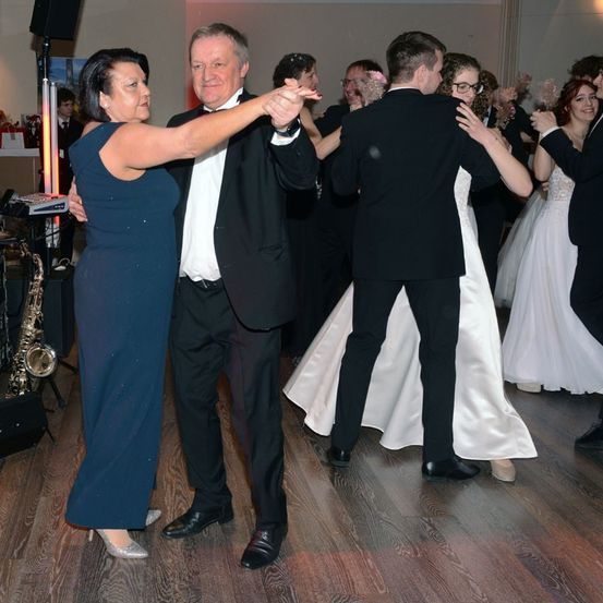 Eine Gruppe von Menschen tanzt auf einem hölzernen Boden bei einer Party. Eine Frau in einem blauen Kleid und ein Mann in einem Smoking führen den Tanz an, während andere folgen. Ein Saxophon und eine Tastatur sind im Hintergrund zu sehen.