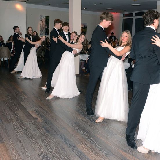Eine Gruppe von Menschen in formeller Kleidung tanzt auf einem Holzboden in einem Saal. Einige sind Paare, während andere in Gruppen tanzen. Der Raum ist gut beleuchtet, mit einigen Stühlen im Hintergrund.