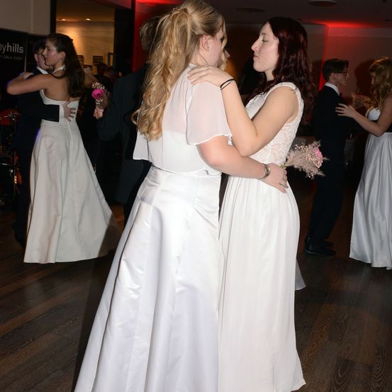 Zwei Frauen in weißen Kleidern tanzen in einem Raum mit anderen Gästen. Eine hält einen rosa Blumenstrauß. Sie haben ihre Arme umeinander geschlungen.