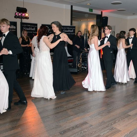 Eine Gruppe junger Menschen in formeller Kleidung tanzt auf einem Holzboden mit einer Scheinwerferbeleuchtung darüber. Ein Saxophon ist im Hintergrund sichtbar.
