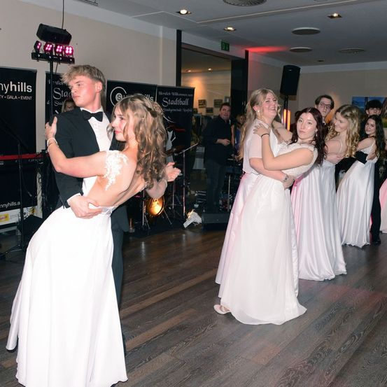 Eine Gruppe von Menschen tanzt in einem Saal auf einem Holzboden. Einige sind in formeller Kleidung. Der Bereich ist gut beleuchtet. Musikinstrumente stehen auf der Bühne. Poster und Lampen sind an der Wand montiert.
