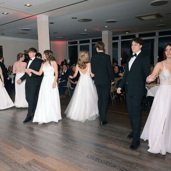 Eine Gruppe von Menschen tanzt bei einer Hochzeitsfeier. Mehrere Männer und Frauen in formeller Kleidung tanzen paarweise auf einem Holzboden.