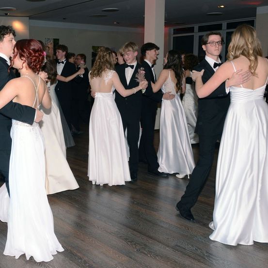 Mehrere Paare in formeller Kleidung tanzen in einem Saal. Die Frauen tragen weiße Kleider und die Männer schwarze Anzüge mit Fliegen. Der Boden ist aus Holz und die Beleuchtung ist gedimmt.