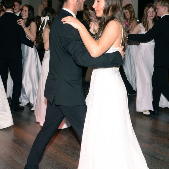 Ein Paar tanzt auf einem hölzernen Boden bei einer formellen Veranstaltung. Die Frau trägt ein weißes Kleid und der Mann einen schwarzen Anzug. Hinter ihnen tanzen auch andere Gäste.