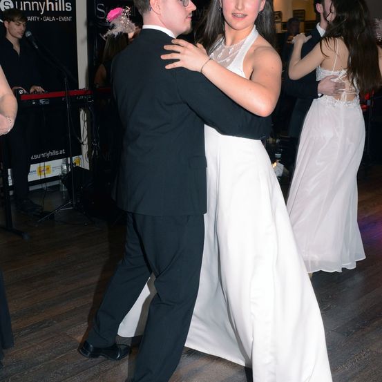 Ein Paar tanzt bei einer formellen Veranstaltung, die Frau in einem weißen Kleid und der Mann in einem schwarzen Anzug. Ein weiteres Paar tanzt hinter ihnen, und ein Musiker spielt ein Keyboard.