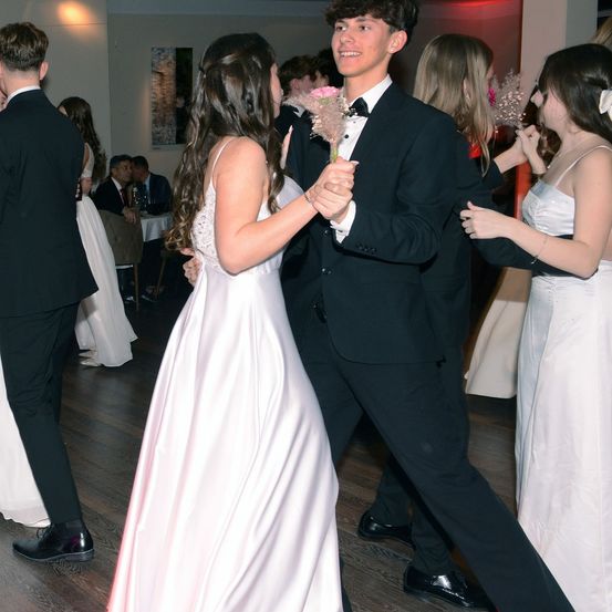 Ein junges Paar tanzt auf einer Tanzfläche, umgeben von anderen Gästen. Der Mann trägt einen Smoking und die Frau ein weißes Kleid. Beide halten rosa Blumen.